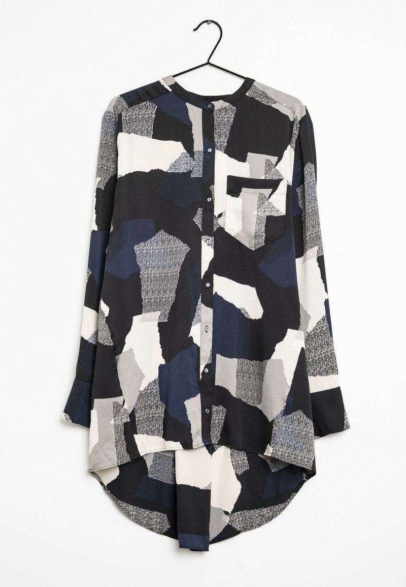Chemise à manches longues boutonnée avec un motif patchwork abstrait irrégulier en noir, blanc, gris et bleu marine, suspendue sur un cintre noir.