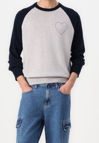 Personne portant un pull gris clair avec des manches raglan noires et un cœur cousu sur la poitrine, assorti à un jean cargo en denim bleu.