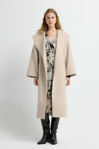 Abrigo de lana beige con solapas anchas, mangas sobredimensionadas y corte recto, superpuesto a un vestido estampado y combinado con botas negras hasta la rodilla.