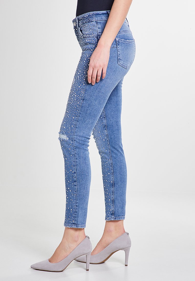 Jeans skinny in denim blu con abbellimenti di piccole perle sui lati, caratterizzati da un design con ginocchio strappato e vita standard.