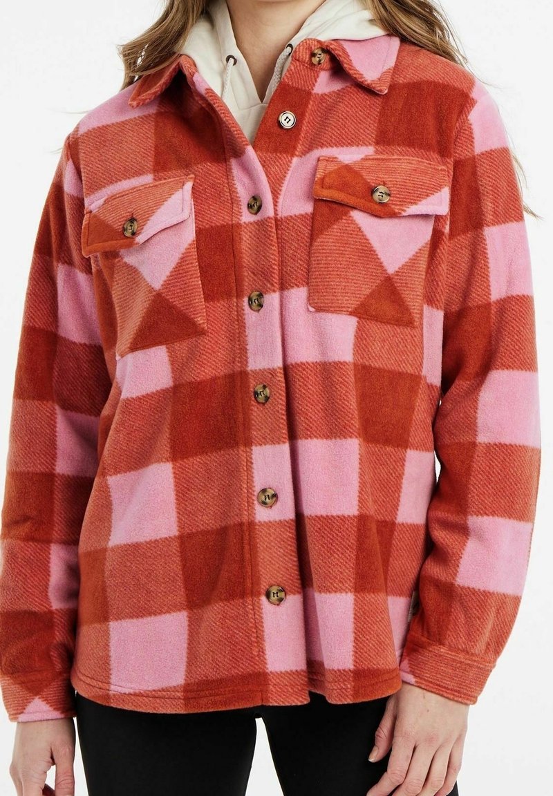 Femme portant une veste à carreaux rouges et roses boutonnée avec deux poches poitrine, par-dessus un sweat à capuche de couleur claire.