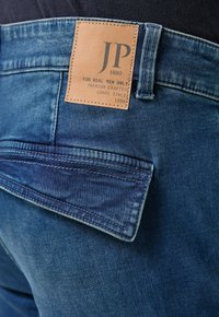 Granatowe dżinsy z denimu z jasnobrązową skórzaną naszywką z logo "JP 1880", widocznym przeszyciem i detalem kieszeni na plecach.