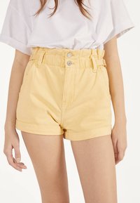Höga gula denimshorts med uppvikta fållar, med framfickor, bälteslipsar och metallknäppningar.