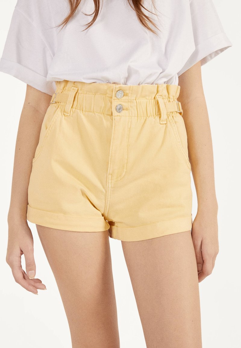 Höga gula denimshorts med uppvikta fållar, med framfickor, bälteslipsar och metallknäppningar.