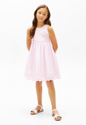 Ung pige stående med hænderne bag ryggen, iført en ærmeløs lyserød kjole med hulmønsterkant og hvide sandaler.