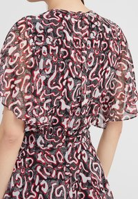 Blouse transparente avec un motif abstrait noir, blanc et rouge. Manches bouffantes avec des poignets élastiques et un détail de fermeture en forme de clé à l'arrière.