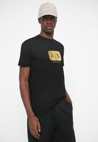 Άνδρας που φοράει μπεζ καπέλο Armani Exchange, μαύρο μπλουζάκι Armani Exchange με χρυσό λογότυπο και μαύρο παντελόνι, με τα χέρια στις τσέπες, σε λευκό φόντο.