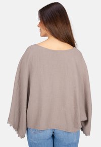 Hellbrauner, oversized, strukturierter Baumwolloberteil mit weiten Ärmeln und gezacktem Saum, getragen mit blauen Jeans, von hinten gezeigt.