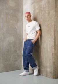 Weißes T-Shirt mit Logo, blaue weite Jeans und weiße Turnschuhe. Glatte Textur, minimalistisches Design und rosa Haarstyle. Graue Betonschwall-Hintergrund.
