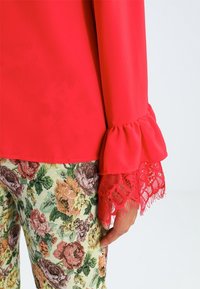 Blusa rossa con maniche a puff in pizzo, abbinata a pantaloni a fantasia floreale nelle tonalità di rosa, verde e giallo su uno sfondo chiaro.