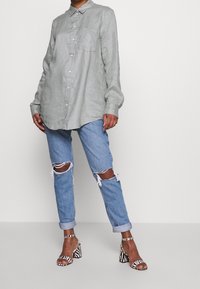 Camisa abotonada de lino verde claro, de corte relajado, con mangas largas y bolsillo en el pecho; combinada con vaqueros azul desgastados y tacones a rayas.