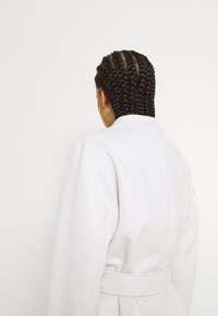 Jasnoszary płaszcz z polaru z paskiem, kołnierzem z wycięciami i długimi rękawami. Widok od tyłu ukazuje splecione włosy. Gładka faktura i minimalistyczne detale.