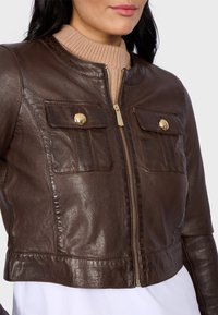 Giacca cropped in pelle marrone con zip frontale, colletto a coste e due tasche sul petto con bottoni dorati. Texture liscia, design aderente.