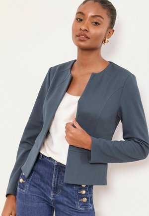 Frau trägt dunkelgraues Blazer mit offenem Schnitt über weißem Oberteil und hoch taillierte blaue Jeans mit dekorativen goldenen Knöpfen, posiert vor weißem Hintergrund.