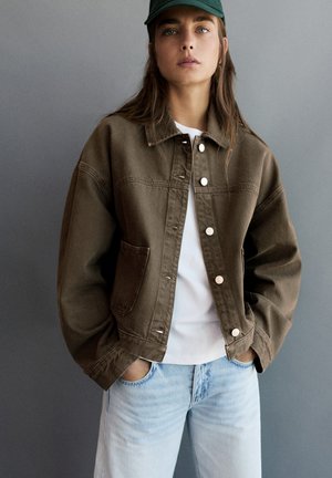 Femme portant une casquette verte, une veste marron boutonnée, une chemise blanche et un jean bleu clair, les mains dans les poches, sur un fond gris.