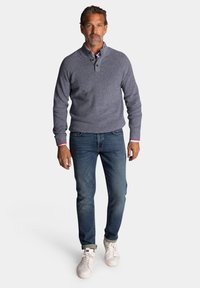 Heren navy gebreide trui met hoge kraag en knoopsluiting met drie knopen, gecombineerd met blauwe jeans en witte sneakers. Casual, comfortabele pasvorm.