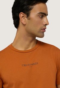 Camiseta de manga corta naranja de algodón, con un cuello redondo y un logo impreso en gris en el pecho. Textura suave.
