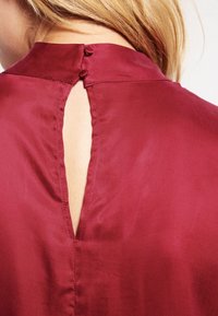 Burgundy silkesblus med hög krage, nyckelhål bak och två knappar i nacken, som visar upp en slät och mjuk textur.