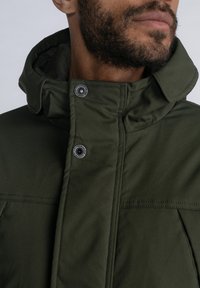 Chaqueta verde oliva con alto cuello, broches metálicos y costuras visibles. La tela tiene una textura suave y un diseño estructurado.