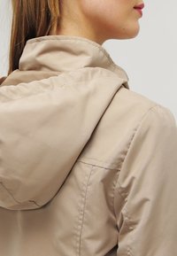 Giacca beige con cappuccio caratterizzata da una texture liscia e morbida e da un colletto sicuro. Dettagli di cucitura ben visibili sulle spalle e nelle tasche.