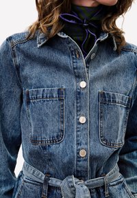 Lichte blauwe denim jurk met een kraaghalslijn, vier voorzakken, zilveren knopen en een stoffen riem in de taille. Textuurafwerking.