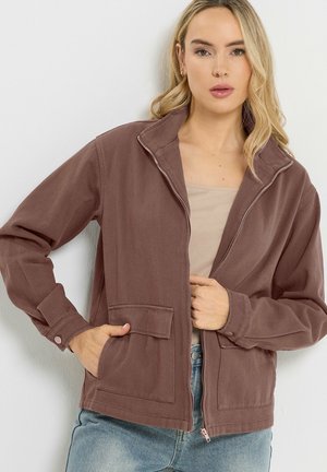 Kobieta w mauve kurtce na zamek, nałożonej na beżowy top i jasnoniebieskie dżinsy, z jedną ręką w kieszeni, a drugą trzymającą kurtkę.