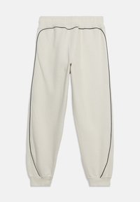 Beige joggebukser med svart piping langs yttersidene på bena, elastisk midjebånd og elastiske mansjetter ved anklene, lagt flatt på hvit bakgrunn.