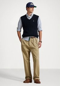 Σκούρο μπλε γιλέκο πλεκτό με καλώδιο πάνω από μπλε ριγέ πουκάμισο, χακί παντελόνι και καφέ παπούτσια. Ολοκληρώνεται με μπλε καπέλο που φέρει λευκό κείμενο.