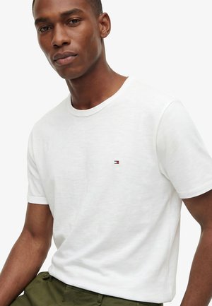 Uomo che indossa una maglietta bianca semplice a maniche corte con un piccolo logo a bandiera sul petto e pantaloni verde oliva su sfondo bianco.