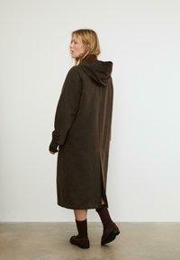 Donna con capelli biondi che indossa un lungo cappotto marrone con cappuccio, calze marroni scure e scarpe marroni, in piedi su un pavimento grigio contro un muro bianco.