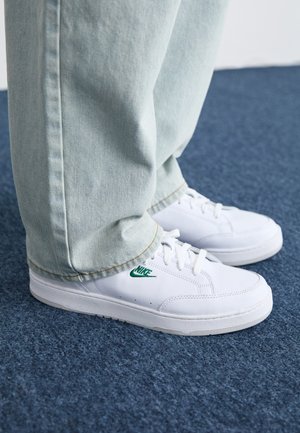 Fehér bőr sportcipők zöld Nike logóval, kerek orral, lapos talppal, világoskék farmernadrággal párosítva, kék szőnyegen bemutatva.