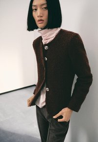 Cardigan marrone, texture lavorata, con tre bottoni lucidi, indossato sopra una maglietta rosa chiaro. Taglio ed eleganza di design con maniche lunghe e scollatura tonda.