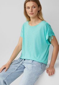 Hellblaues kurzärmeliges Oberteil mit rundem Halsausschnitt, das mit gerafften Details an den Ärmeln versehen ist. Kombiniert mit hellblauen Jeans.