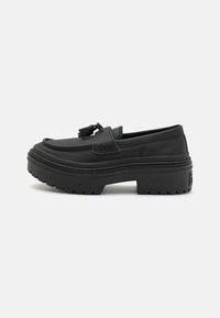Svart plattform loafers laget av glatt skinn, med rund tå, dekorative dusker og en tykk gummisåle med strukturert mønster.