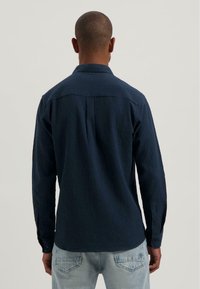 Man met kort haar, gekleed in een textuur donkerblauw langslavend shirt en lichtblauwe broek, staand met zijn gezicht weg van de camera tegen een effen achtergrond.
