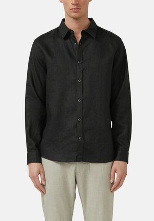 Mann, der ein schwarzes langärmeliges Hemd mit Knöpfen und hellbeige Hosen trägt, steht mit entspannten Armen vor einem schlichten Hintergrund.