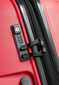 Red hard-shell suitcase with black dual zipper pulls locked into a built-in TSA-approved combination lock.
