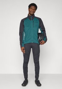 Sportoutfit für Herren mit einer türkisfarbenen und schwarzen Reißverschlussjacke mit hohem Kragen sowie schwarzen Radsportleggins. Zubehör umfasst einen Fahrradhelm.