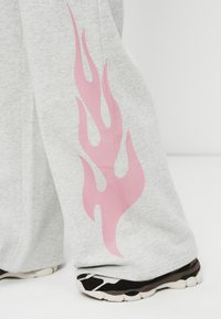 Ανοιχτό γκρι παντελόνια με φαρδιές ίσιες και ένα ροζ φλόγιστρο στο πλάι, φορεμένα με ασπρόμαυρα αθλητικά παπούτσια σε λευκό πάτωμα.