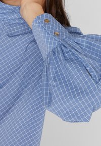 Chemise à carreaux bleus avec une manche structurée froncée, deux boutons décoratifs au poignet et une texture de tissu léger.