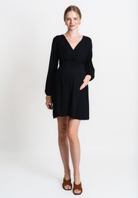 Abito da gravidanza nero in jersey con maniche lunghe, scollo a V e vita arricciata. Indossato con sandali marroni con cinturini. Tessuto morbido, lunghezza al ginocchio.