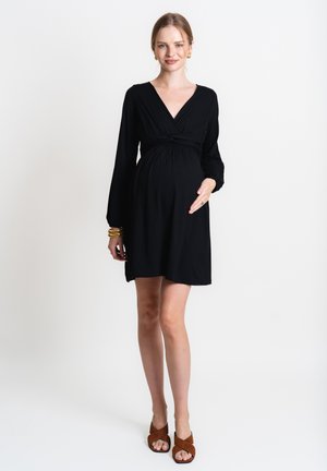 Schwarzes, Jerseykleid für Schwangere mit langen Ärmeln, V-Ausschnitt und gerafftem Bund. Getragen mit braunen, Riemchensandalen. Glatte Textur, knielang.