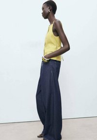 Top giallo a coste senza maniche con taglio dritto e lacci laterali, abbinato a jeans a gamba larga in denim scuro con cuciture in vista e zip.