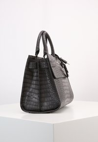 Czarna torebka na ramię z teksturą krokodyla, o wyraźnej formie, z podwójnymi uchwytami i klapką z przodu, wyeksponowana na białym piedestale na jednolitym tle.