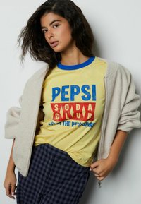 Geel T-shirt met blauwe en rode grafische tekst onder een licht beige fleecejack. Marineblauwe geruite broek maakt de outfit compleet.