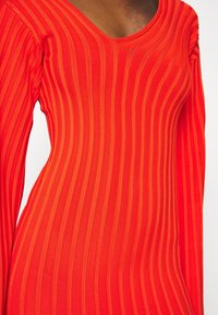 Robe rouge côtelée avec un col rond, des manches longues et des rayures verticales. Le tissu est lisse et élastique.