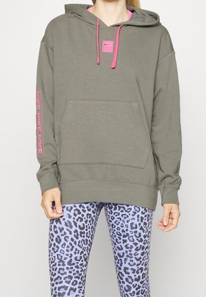 Kobieta w oliwkowej bluzie z kapturem Nike z różowymi sznurkami i logo na rękawie, w towarzystwie fioletowo-czarnych legginsów w panterkę.