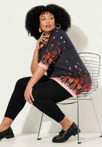 Schwarzes Tunika-Oberteil mit orangefarbenem Blumenmuster, Dreiviertelärmel und pinkem Saum, kombiniert mit schwarzen Leggings und klobigen schwarzen Schuhen.