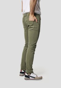 Obcisłe, oliwkowozielone dżinsy z pięcioma kieszeniami, przetarciami oraz brązową skórzaną łatką z logo. Połączone z wielokolorowymi sneakersami.