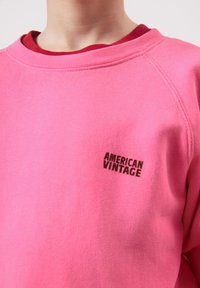 Person trägt einen rosa Pullover mit dem schwarzen Aufdruck "American Vintage" auf der Brust, darunter ein rotes Hemd.